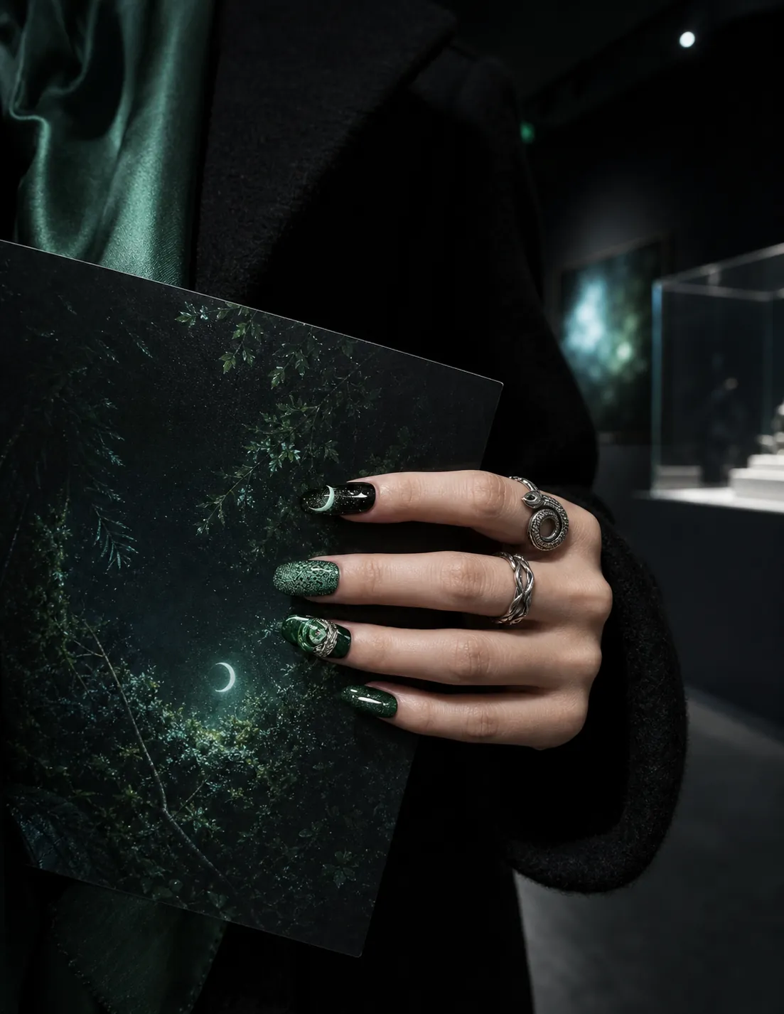 A hand wearing deep green snake-themed nails holds a gallery catalog at night